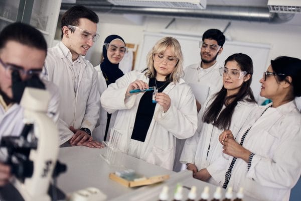 Ett stort gang elever star med labbrockar och samlas kring ett experiment.